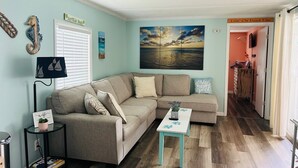 A living room with a sectional sofa, a small table, a bookshelf, and a painting of a beach scene. - 272 Flamingo 2 Bedroom 2 Full Baths (Surfside Beach)
