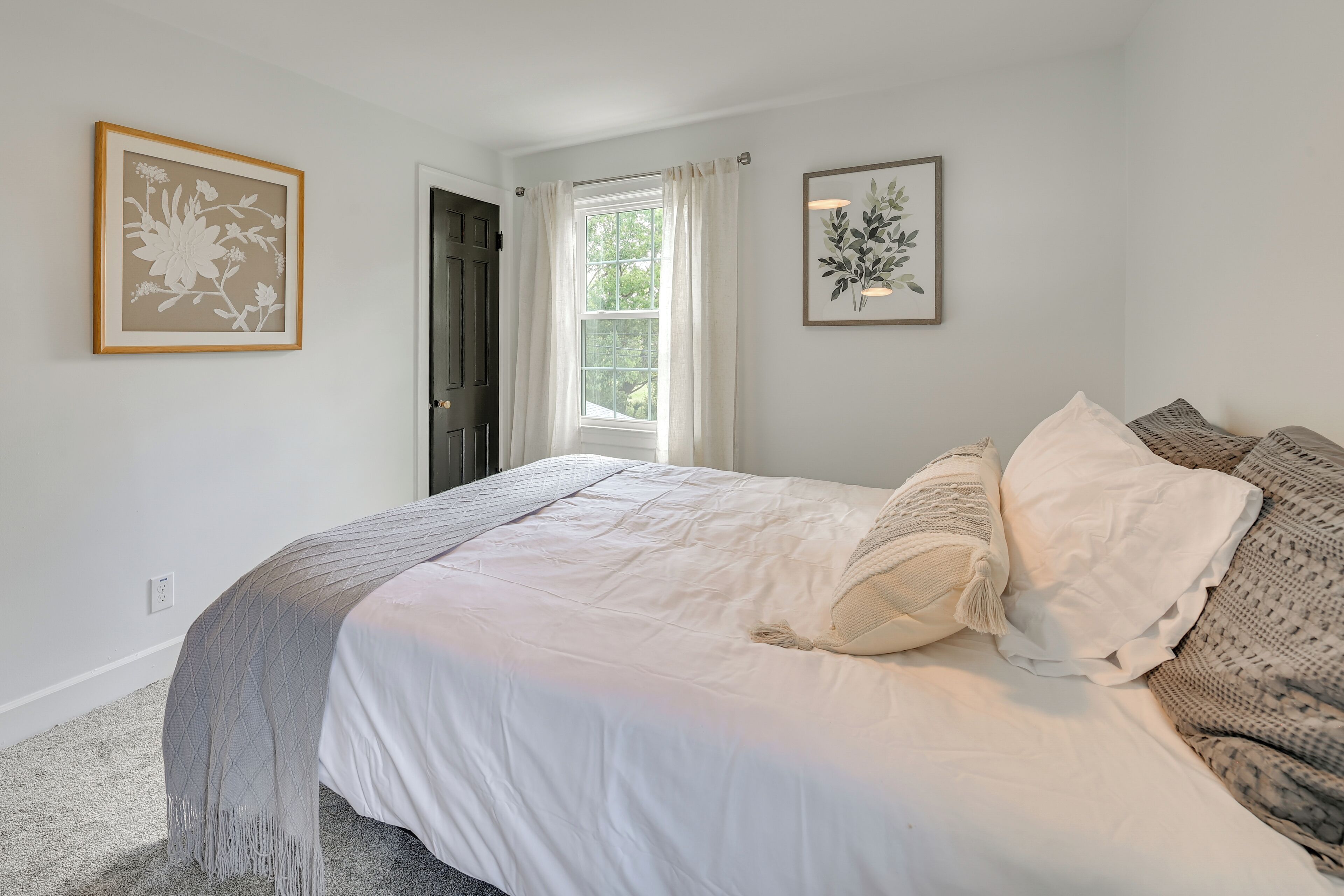 Ein Schlafzimmer mit einem großen Bett, zwei gerahmten Blumenbildern, einem Fenster mit Vorhängen und einer Tür.