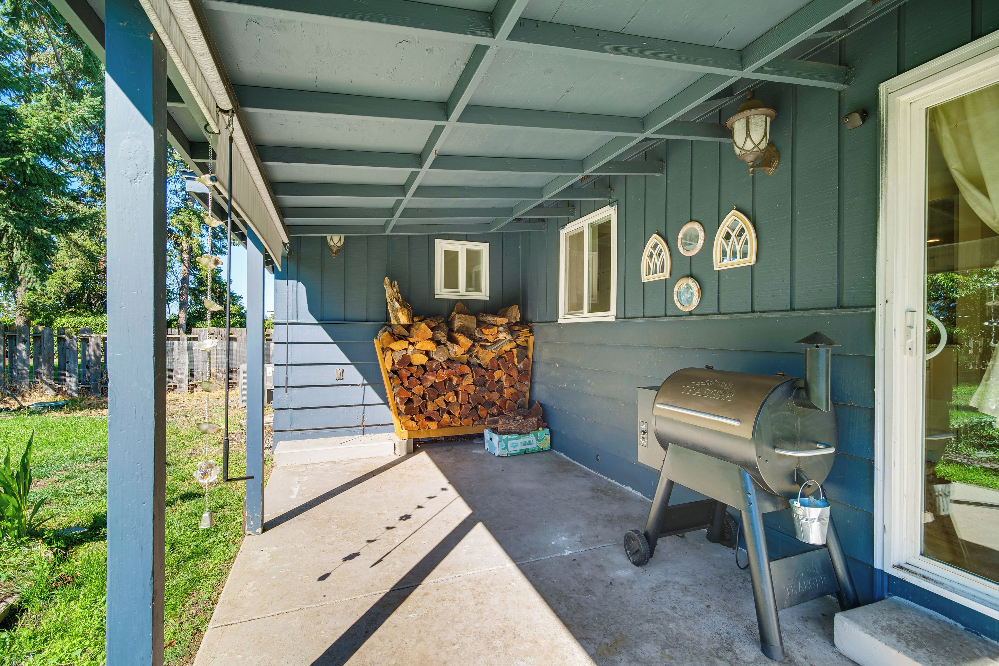 A covered outdoor area with a grill, stacked firewood, and a concrete path leading to a door.