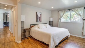 Une chambre avec un grand lit, un plancher en bois, une table de chevet, une commode, une fenêtre avec des rideaux et un tableau accroché au mur.