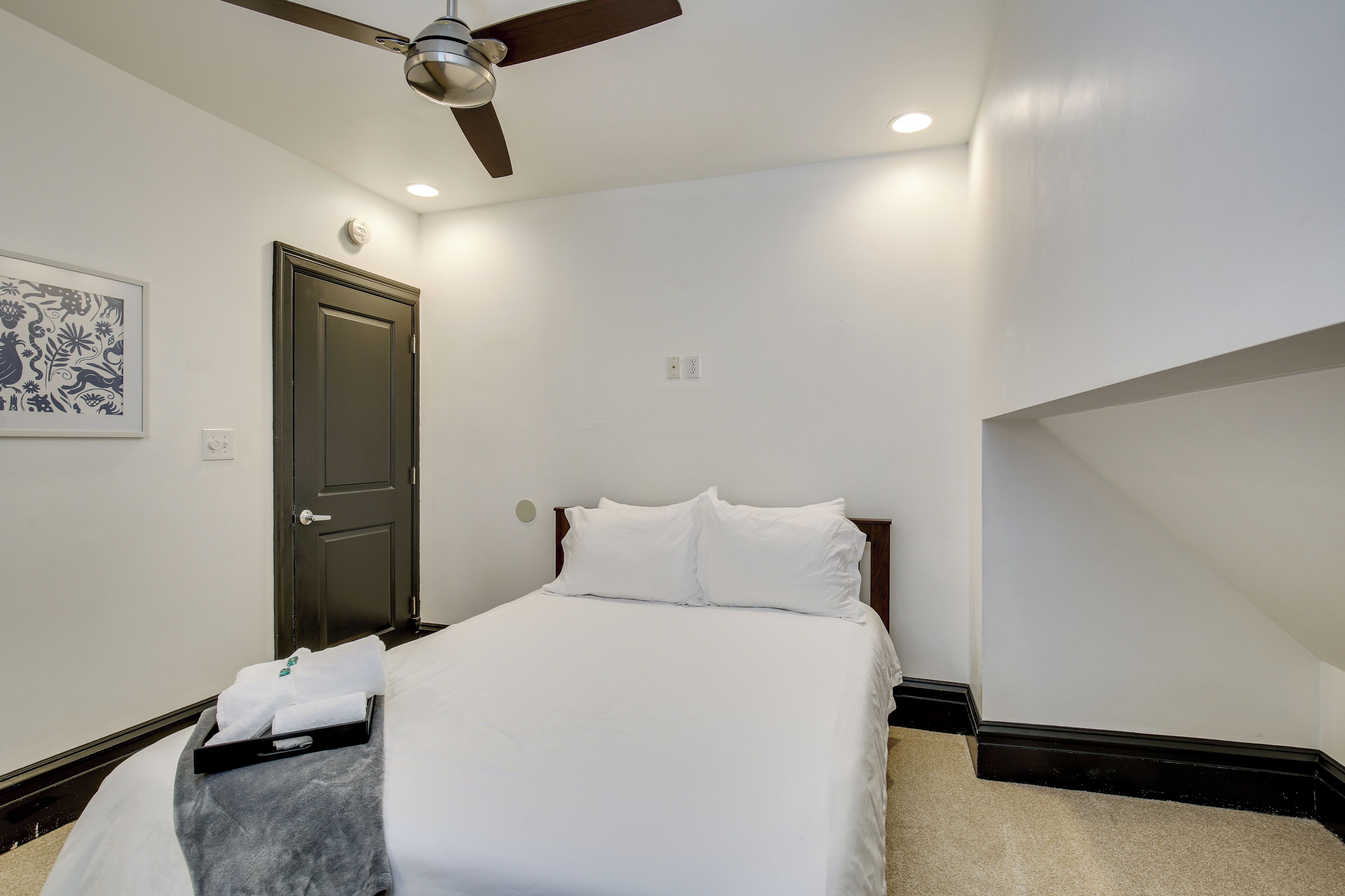Une chambre bien rangée, avec un lit, un ventilateur de plafond, une porte et un luminaire fixé au mur.