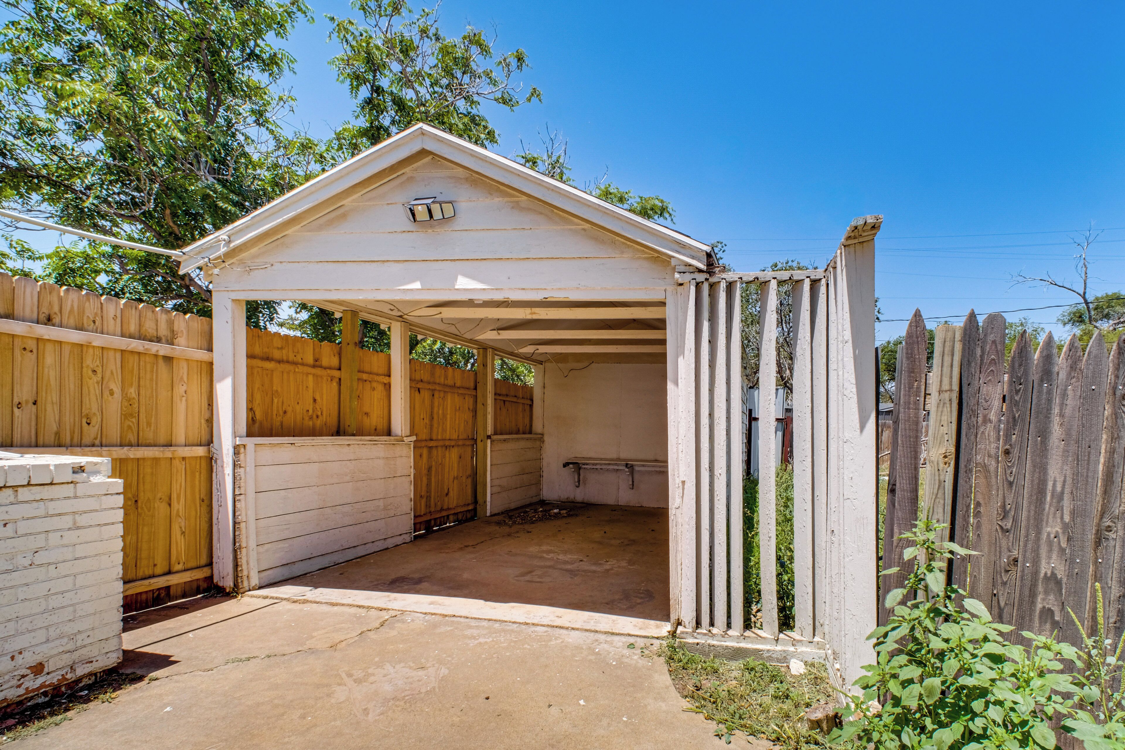 A covered outdoor area with a wooden fence, a concrete floor, and a small shed-like structure with a light fixture.