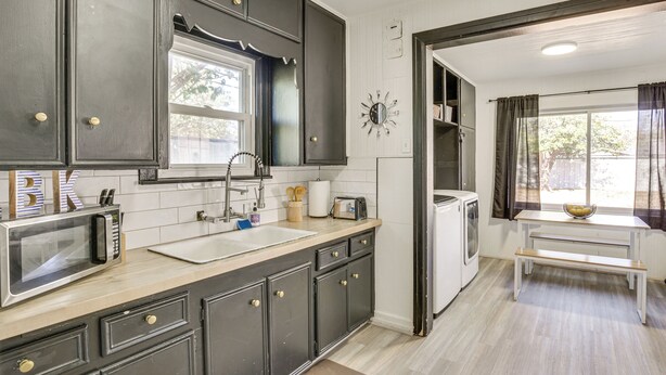 A kitchen with dark cabinets, a microwave, a sink, and a window with curtains. - Time Machine: Games Galore in Lubbock Home By Ttu! (Lubbock)