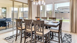 Un coin repas avec une table dressée pour six personnes, entourée de chaises, d’un lustre et de grandes fenêtres ornées de rideaux.