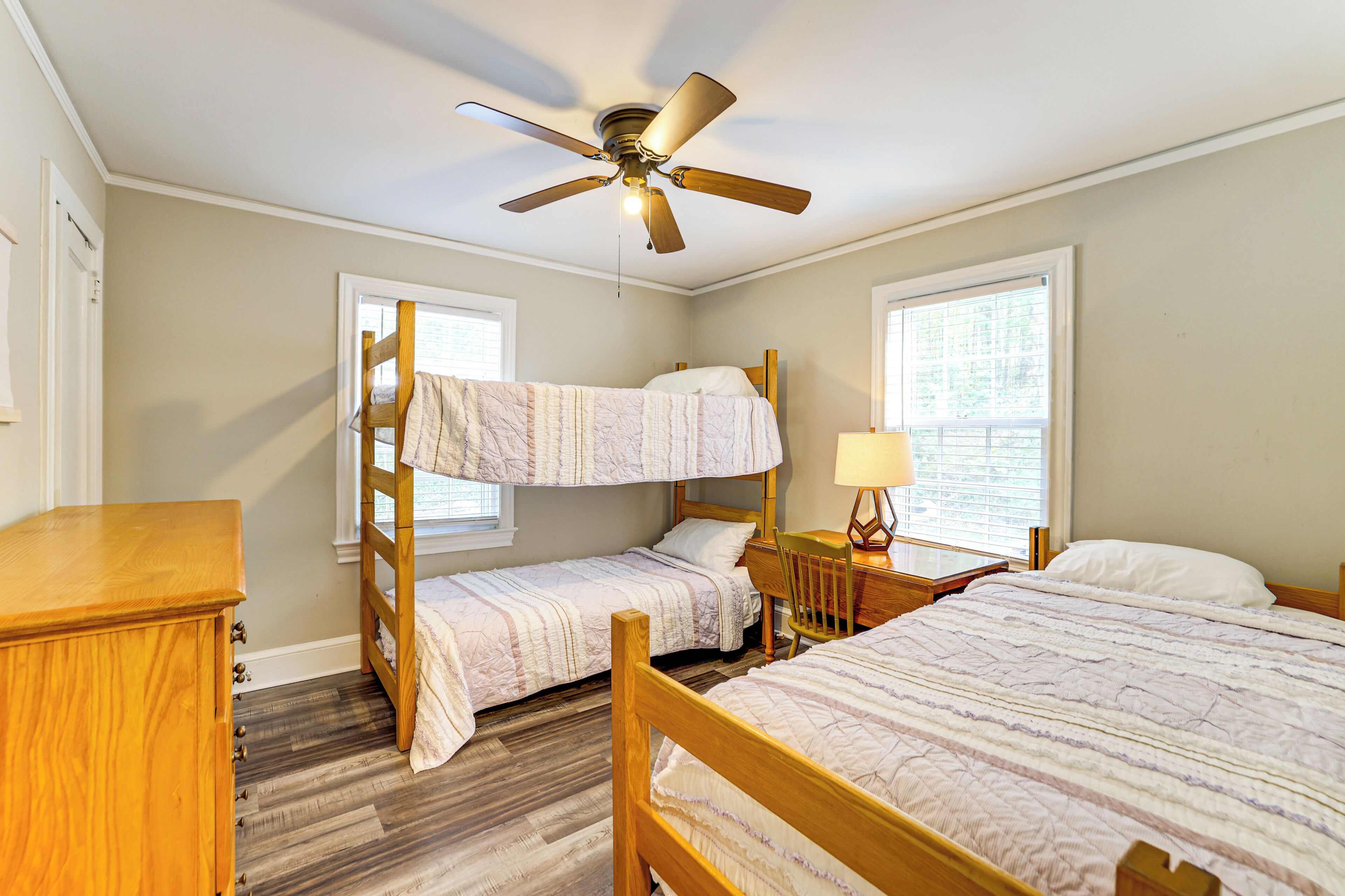 Une chambre avec des lits superposés, un ventilateur de plafond, une commode en bois et deux fenêtres.
