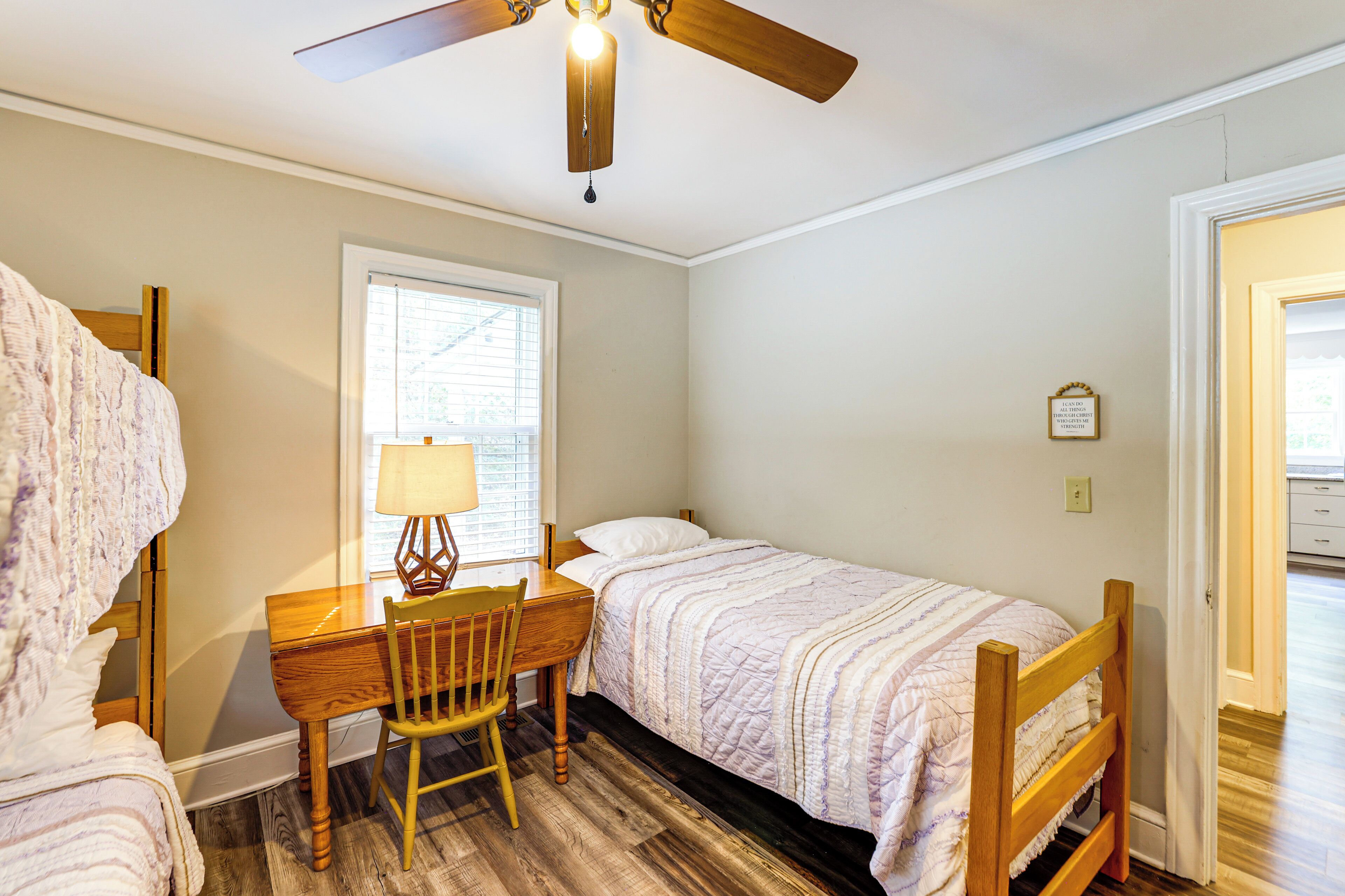 Une chambre avec deux lits simples, un bureau en bois avec une chaise, un ventilateur de plafond et une fenêtre avec des stores.