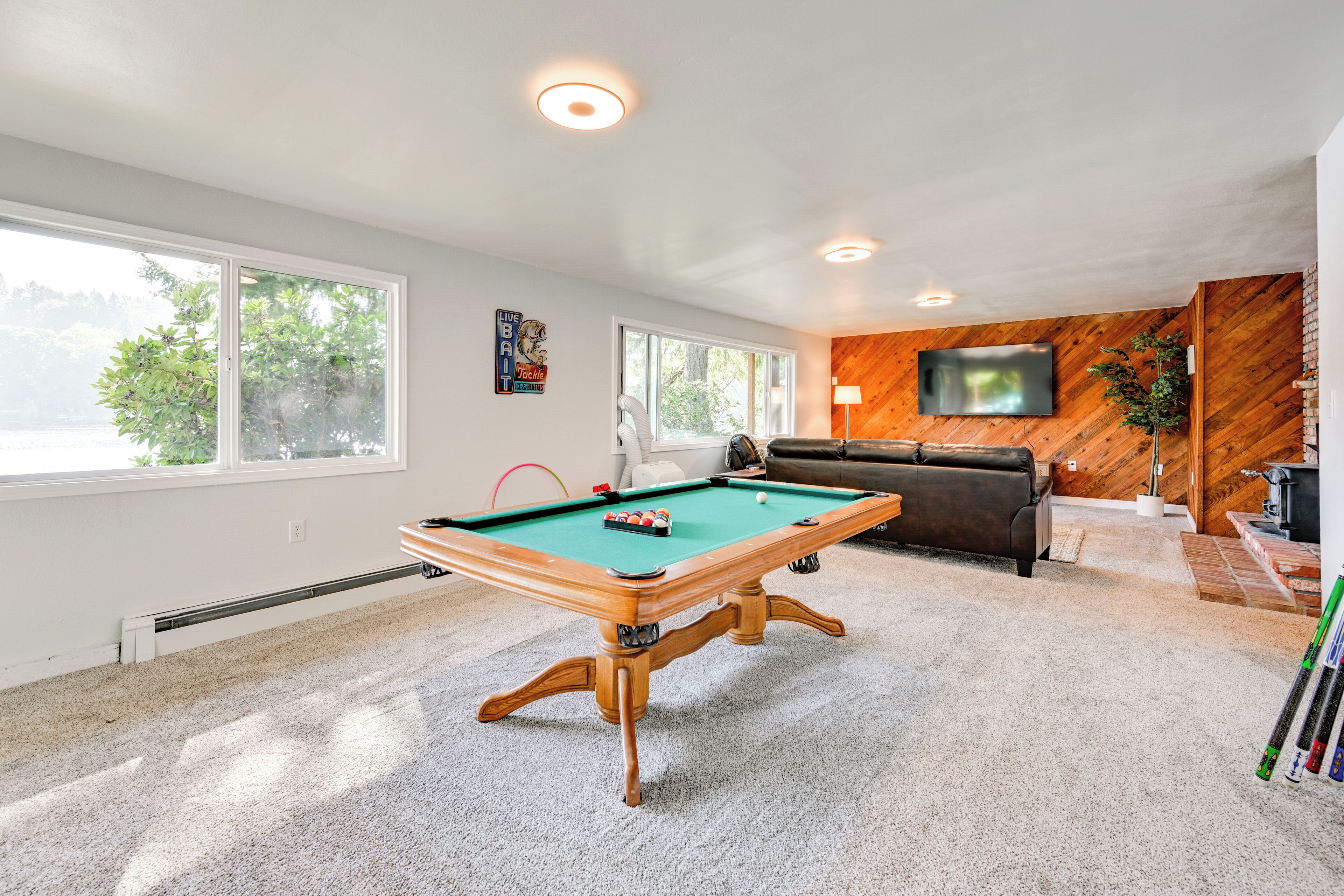 A pool table in a well-lit room with large windows, a flat-screen TV on the wall, and a cozy seating area.