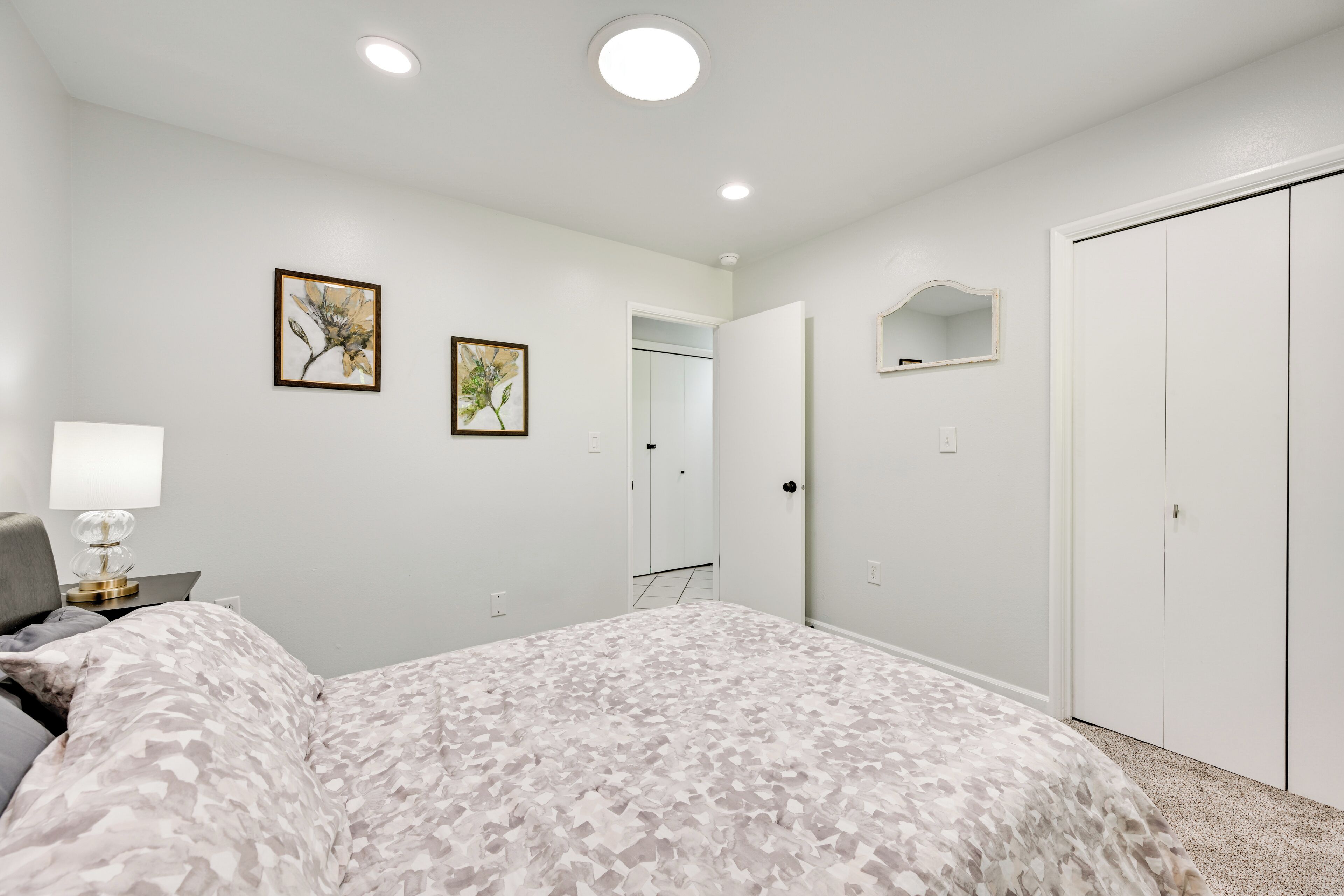 Um quarto com uma cama, uma mesa de cabeceira, um abajur, um guarda-roupa e duas fotos emolduradas na parede.