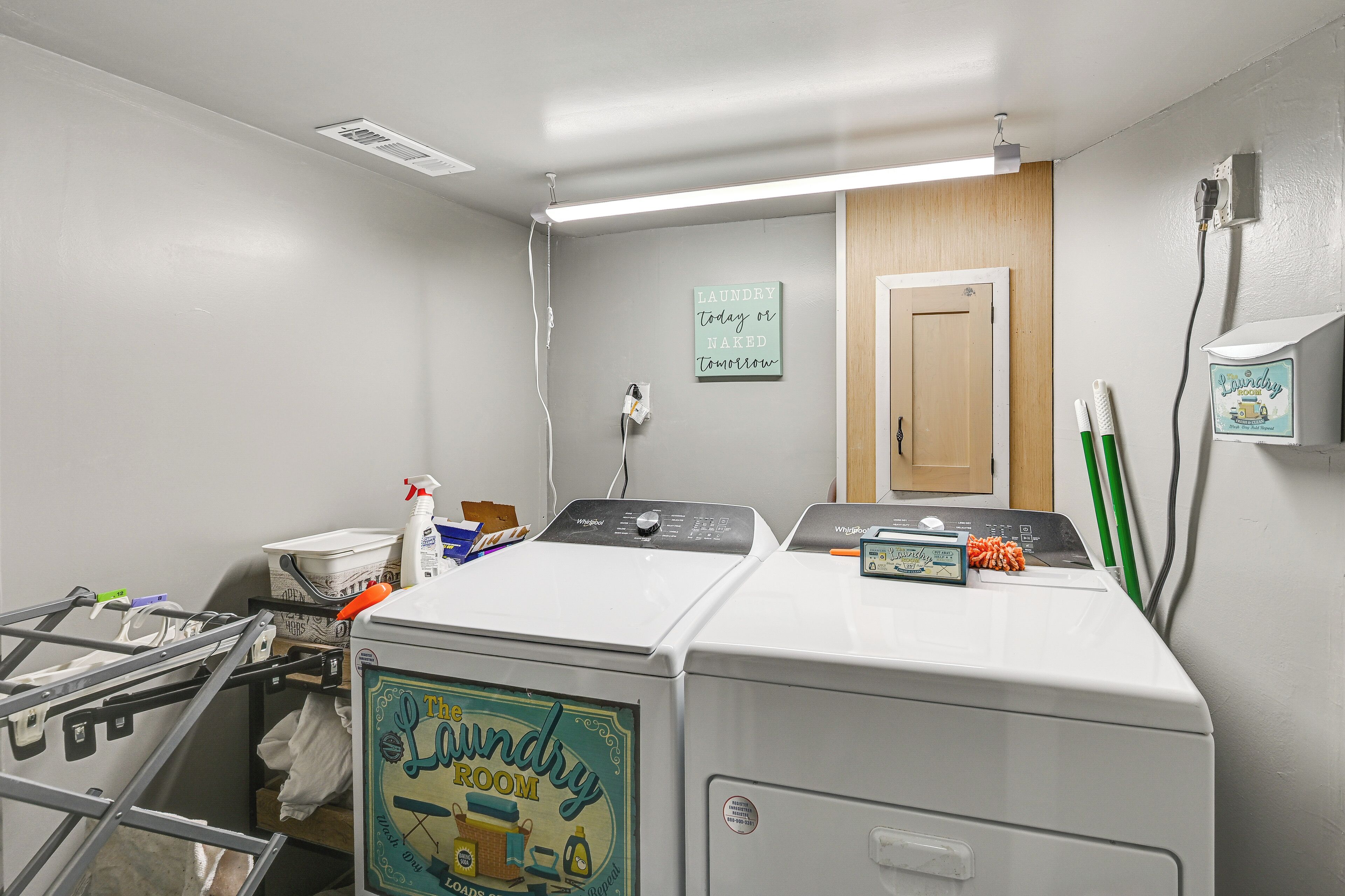 A laundry room with two front-loading washers, a drying rack, a wall-mounted sign, and a door.