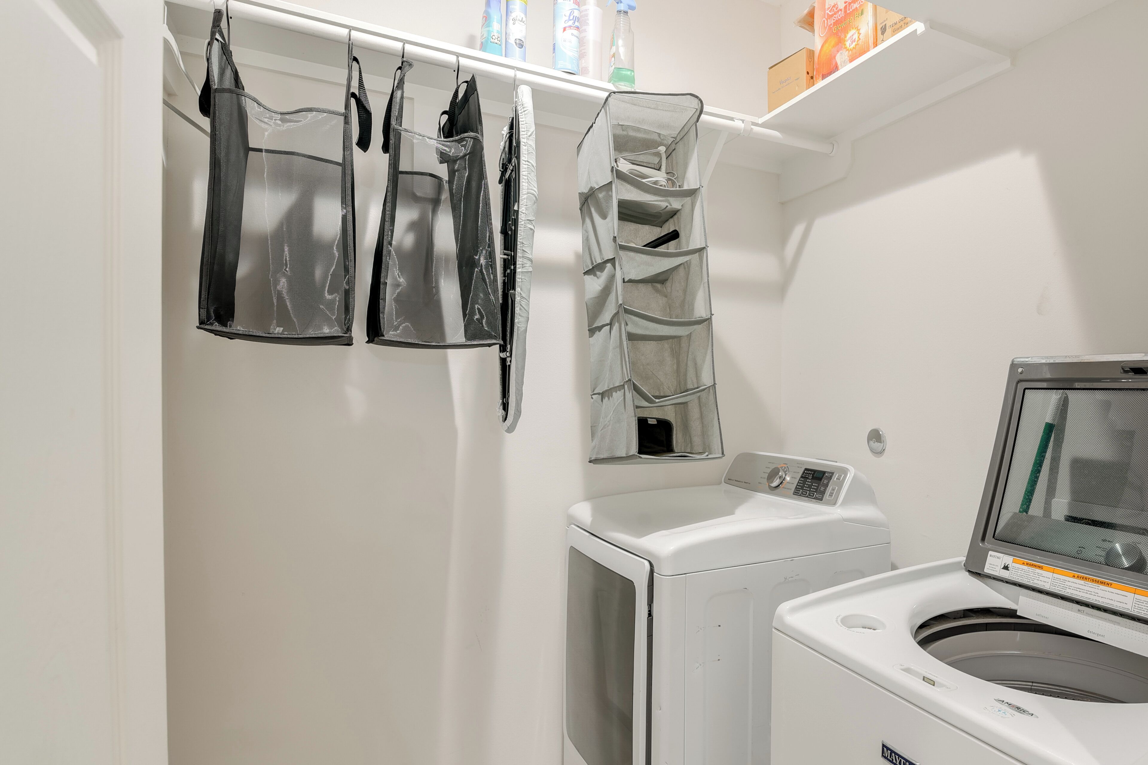 A laundry area with a front-loading washer and dryer, a drying rack with several bags, and a shelf with cleaning supplies.