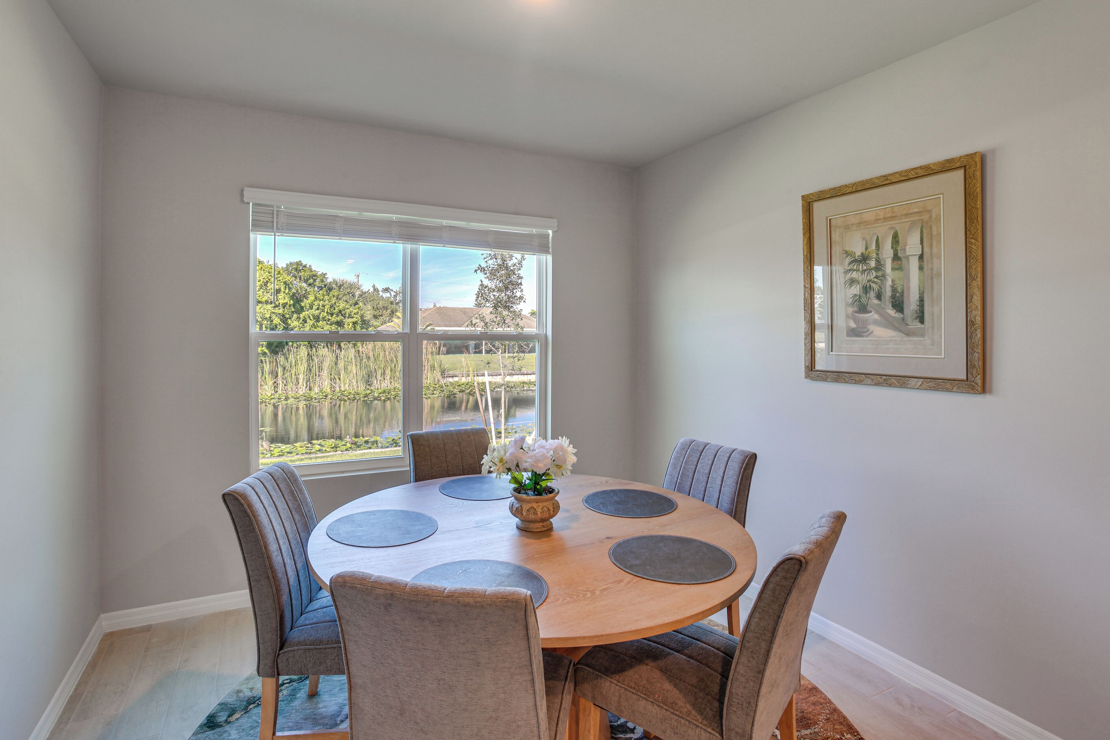 A dining area with a round table, six chairs, a vase with flowers, and a window with blinds.