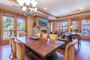 A spacious dining area with a large wooden table, surrounded by upholstered chairs, a stone fireplace, and a view of a mountainous landscape through large windows.