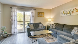 Een moderne woonkamer met twee grijze banken, een glazen salontafel en een eettafel met stoelen. Er is een balkon met uitzicht op bomen en een kunstwerk van een fiets aan de muur.