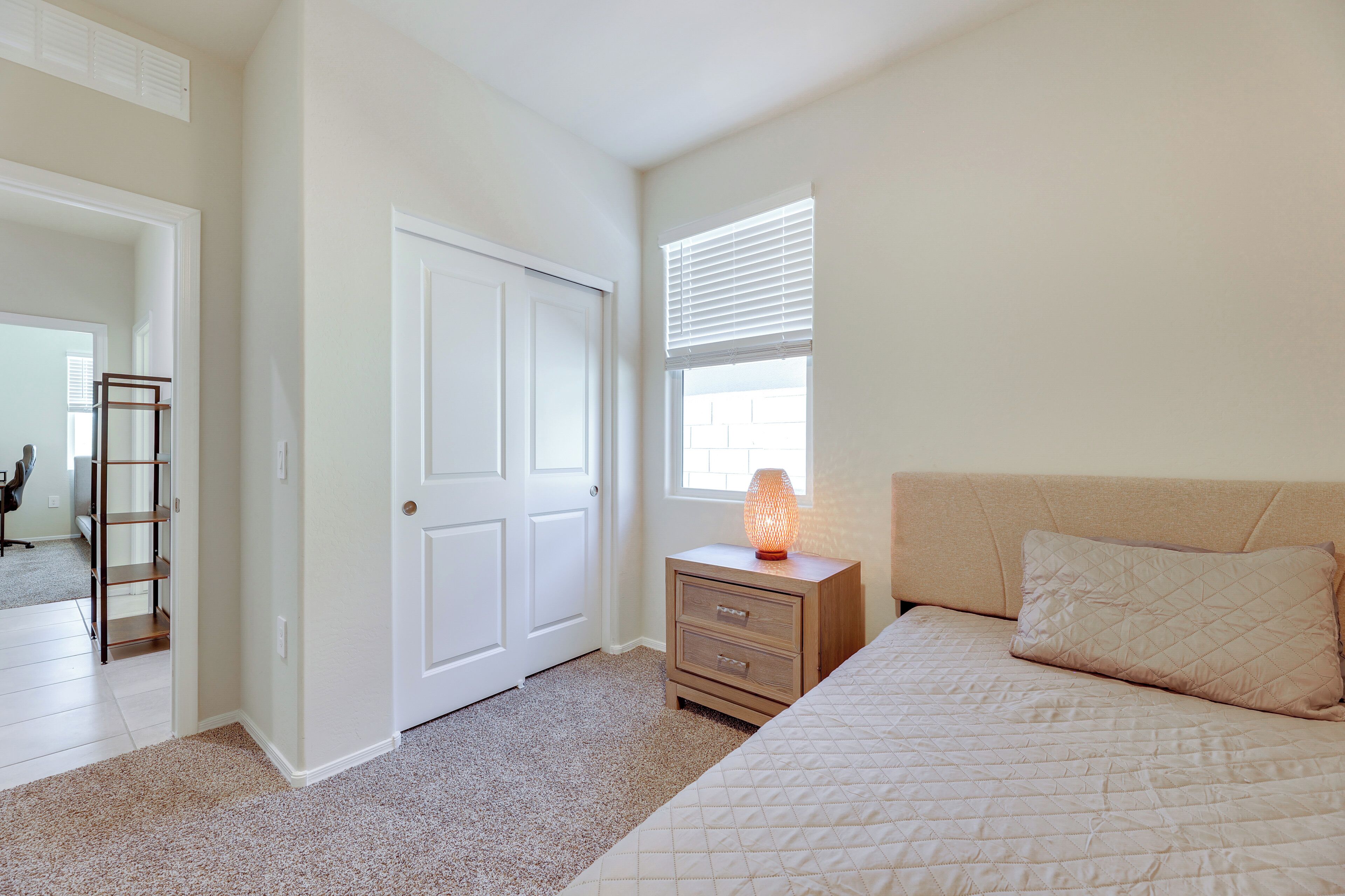 A bedroom with a bed, a nightstand with a lamp, a door, and a window with blinds.