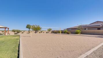 A volleyball court with a net, surrounded by a sandy area, a building with a tiled roof, and sparse vegetation.