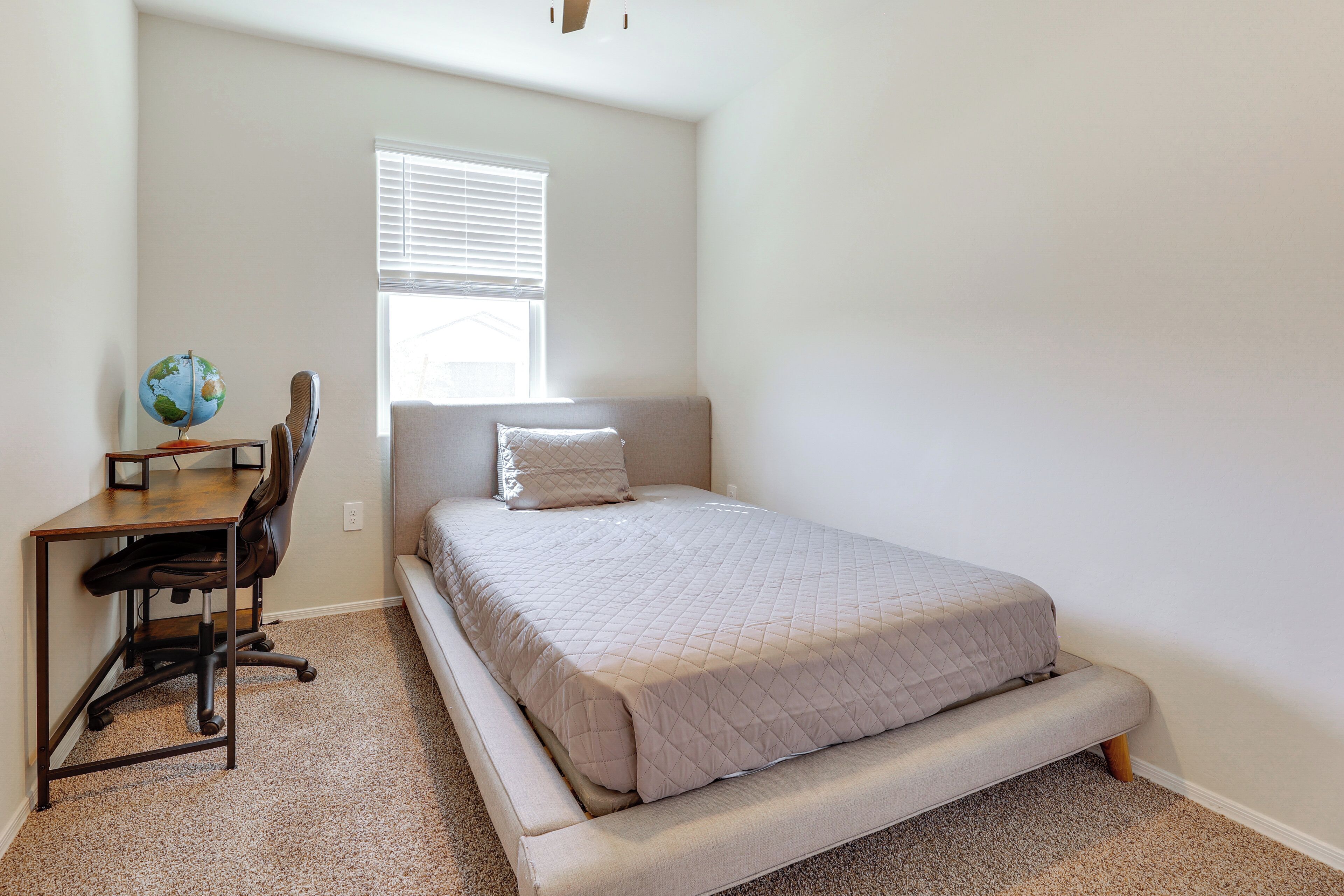 A bedroom with a bed, a desk with a chair, a globe, and a window with blinds.
