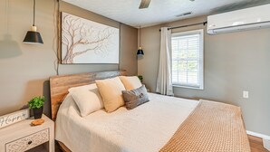Ein Schlafzimmer mit Bett, Nachttisch, Wandbild, Deckenventilator und Fenster mit Vorhängen.