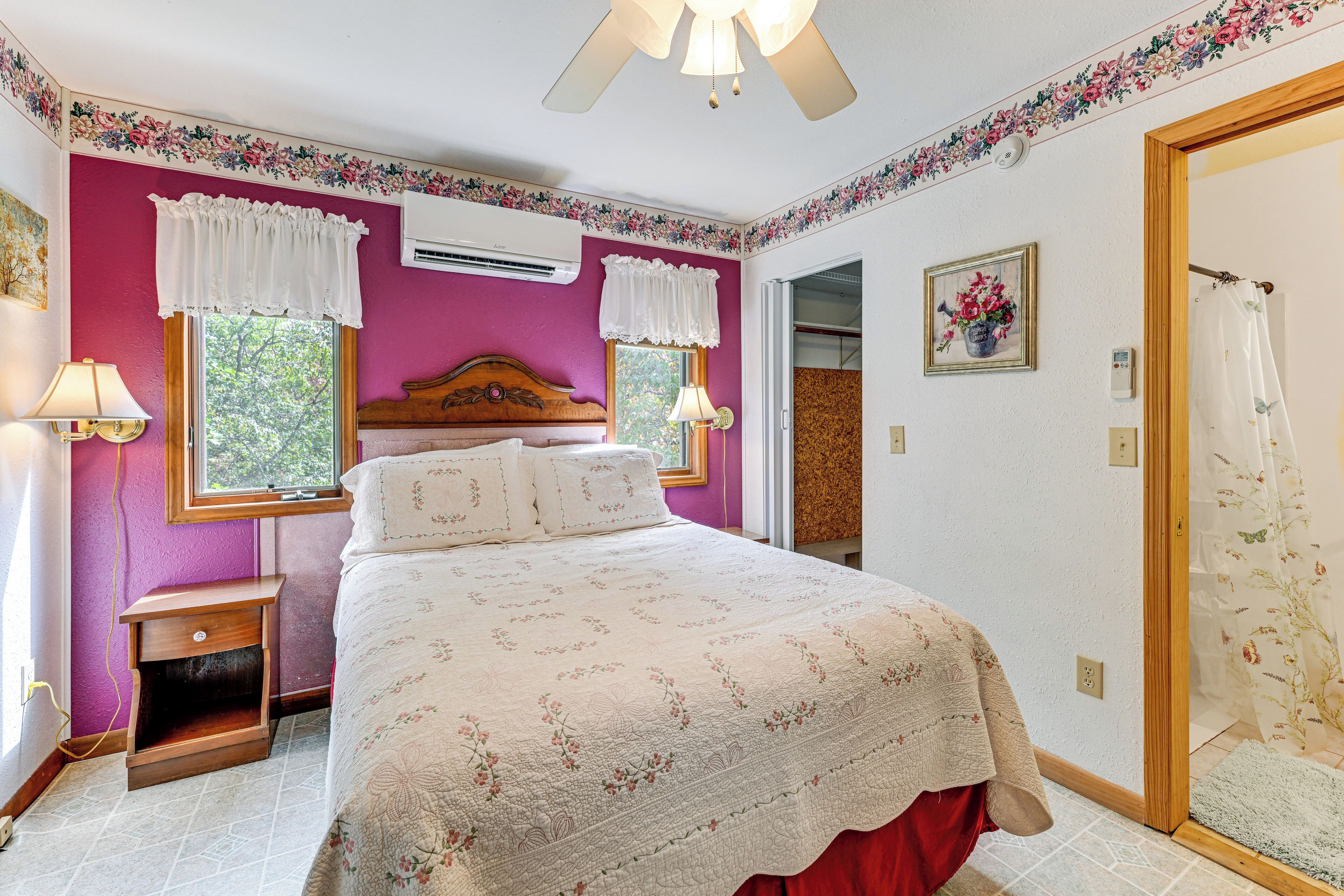 A bedroom with a bed, a nightstand, a window with curtains, a ceiling fan, and a door leading to another room.