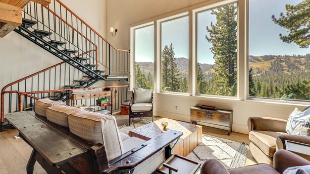 Un vaste espace de vie comprenant un banc en bois, un canapé et un fauteuil. Un escalier avec des rampes en métal mène à un niveau supérieur. De grandes fenêtres offrent une vue sur un paysage boisé.