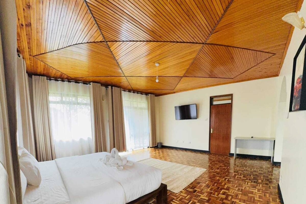 Une chambre d’hôtel avec un plafond en bois, un lit recouvert de draps blancs, un parquet en bois, un téléviseur à écran plat fixé au mur et une porte.
