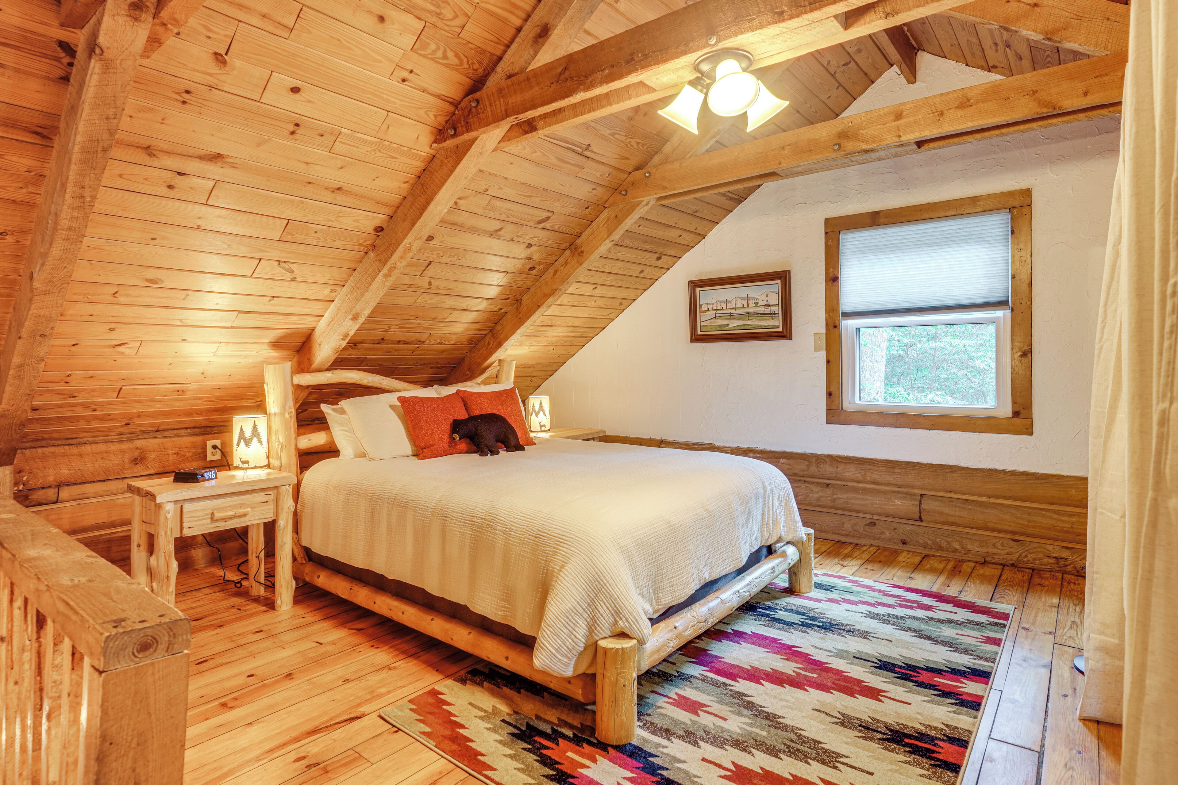 Un dormitorio acogedor en el ático con techo de madera, una cama con un tapete colorido, una mesita de noche con una lámpara y una ventana con persianas.