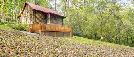 A log cabin with a balcony, surrounded by a forested area with fallen leaves.