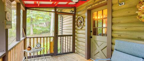 A cabin with a wooden exterior, a porch with a bench, a door with a wheel, and a window with a view of greenery.