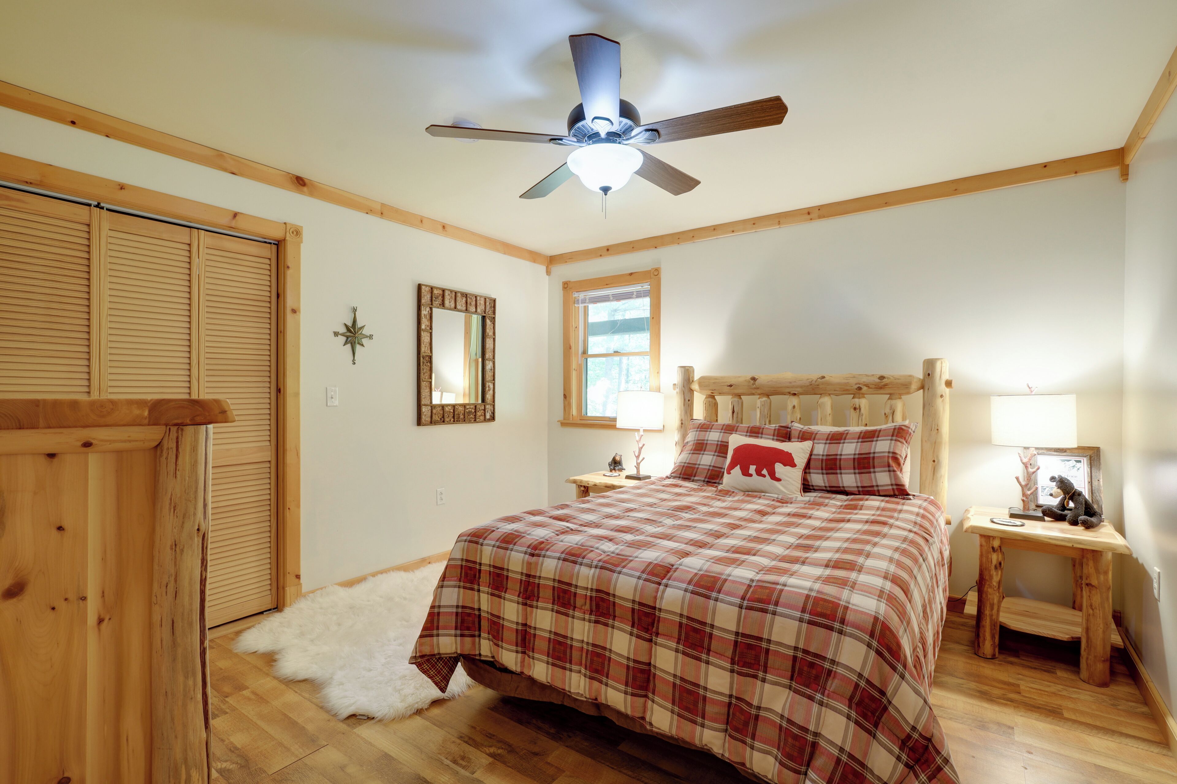 A bedroom with a plaid bedspread, wooden headboard, a ceiling fan, a window, and a wooden nightstand with a lamp.