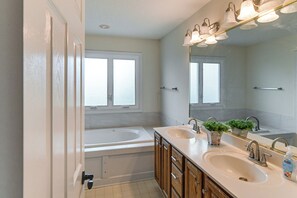 A bathroom with two sinks, a bathtub, wooden cabinets, and a large mirror with lights. - Des Moines Suburb Stay w/ Spacious Layout & Deck! (Johnston)