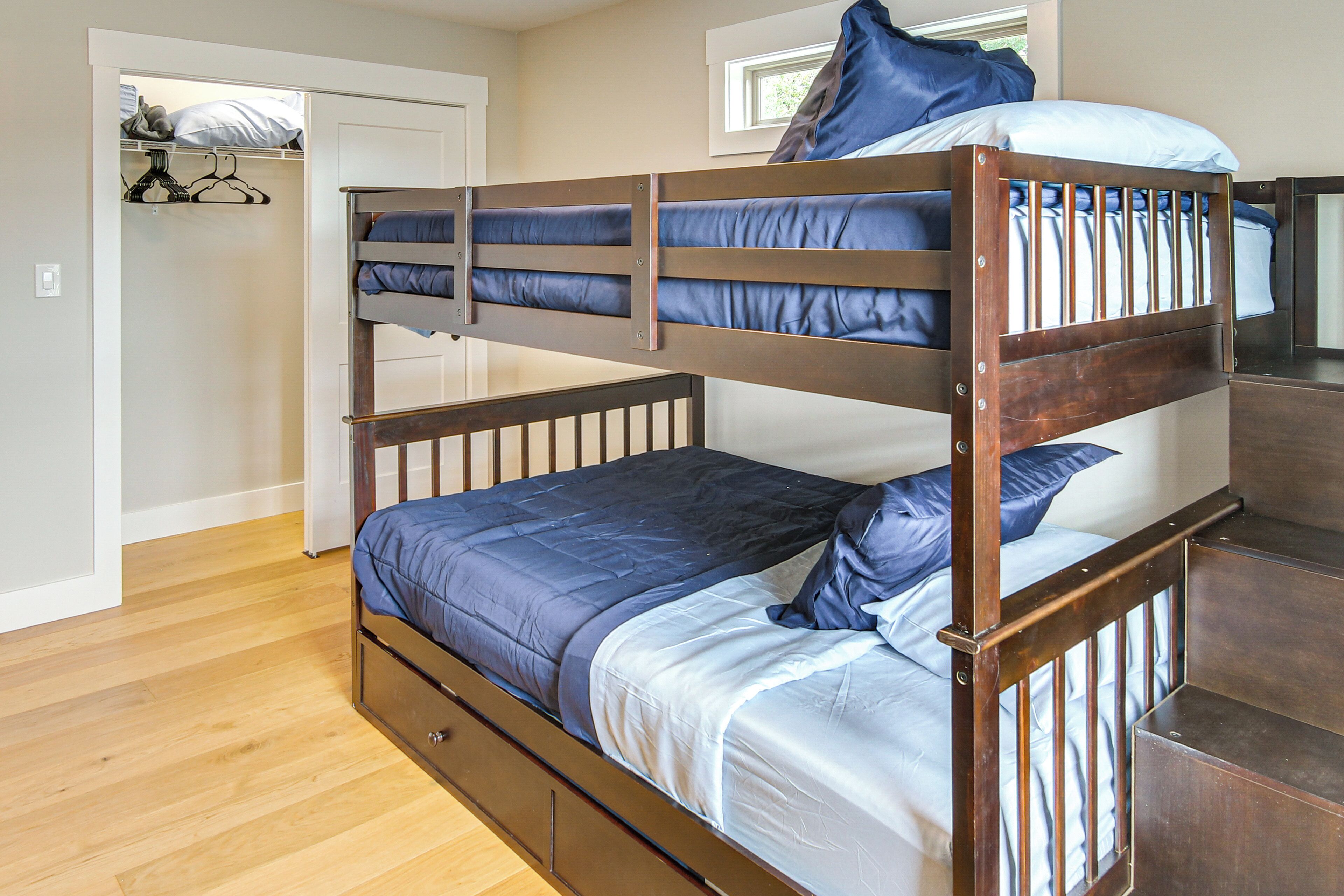 Ein Stockbett mit Holzrahmen und blauer Bettwäsche. Ein Kleiderschrank mit Kleidung und ein Fenster über dem Bett sind ebenfalls vorhanden.