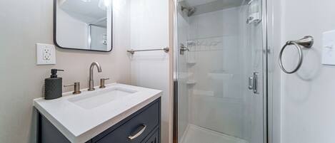 Un baño moderno con ducha envuelta en vidrio, un lavabo blanco con encimera de mármol y un espejo grande sobre el mueble.