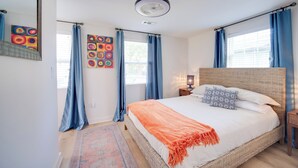 Une chambre avec un grand lit, un ventilateur de plafond et deux fenêtres avec des rideaux bleus.