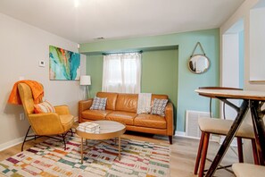 A living room with a brown leather sofa, a round coffee table, a colorful rug, a yellow armchair, and a dining table with chairs.