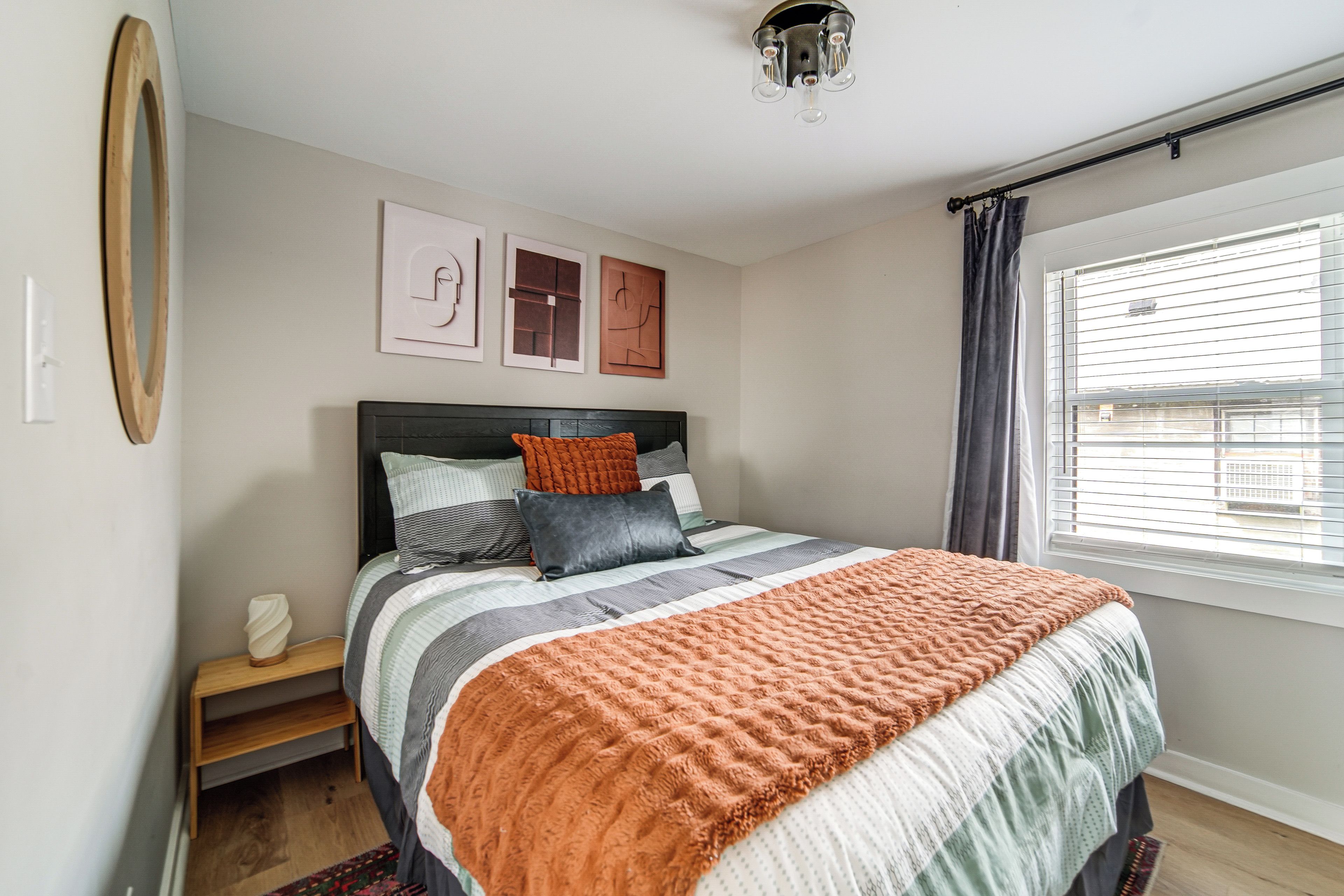A bedroom with a bed, pillows, a bedside table, a lamp, and a window with blinds.