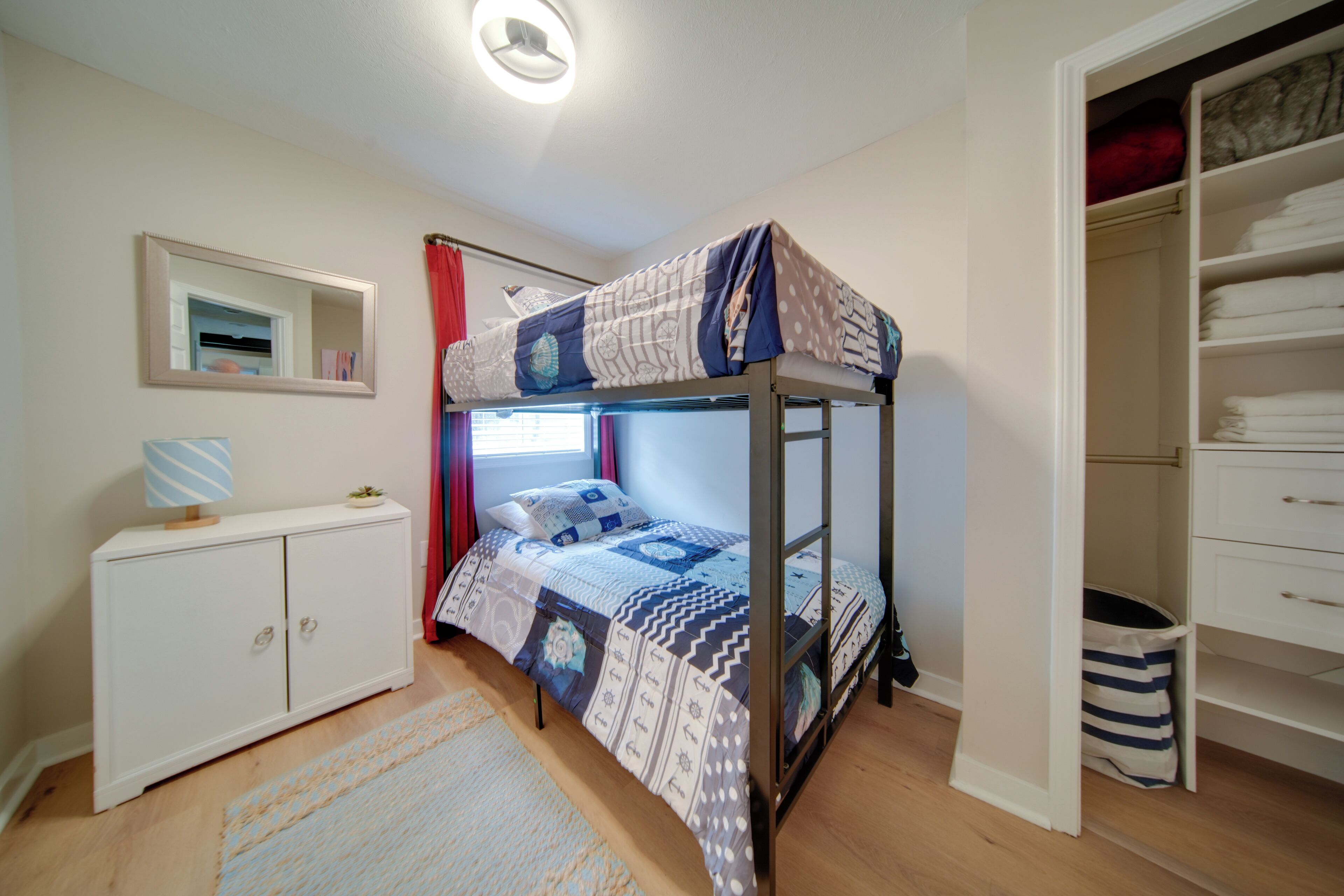A bunk bed with a patterned bedspread, a white dresser with a mirror, and a walk-in closet with folded clothes.