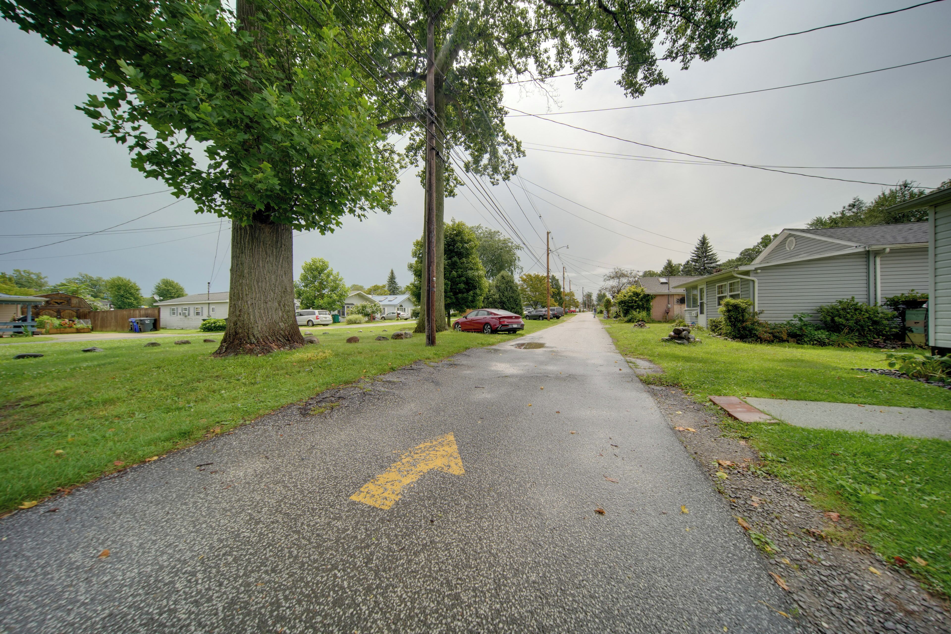 A residential street with houses on both sides, a clear road with a yellow directional arrow, and utility poles with overhead wires.