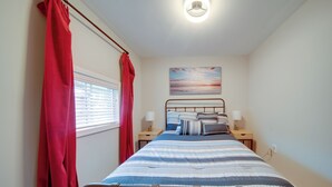 A bedroom with a bed, two bedside tables, a window with blinds, a red curtain, a wall-mounted light fixture, and a painting above the bed. - 'wishin & Fishin' Walkable Erie Home By the Beach! (Erie)