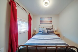 A bedroom with a bed, two bedside tables, a window with blinds, a red curtain, a wall-mounted light fixture, and a painting above the bed. - 'wishin & Fishin' Walkable Erie Home By the Beach! (Erie)
