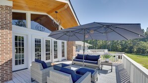 A covered outdoor deck with wicker furniture, a large umbrella, and a view of a brick building. - Game Room & Pool! Luxe Louisburg Farmhouse (Louisburg)