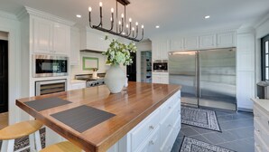 A modern kitchen with white cabinetry, a large wooden island, stainless steel appliances, and a decorative candelabra. - Game Room & Pool! Luxe Louisburg Farmhouse (Louisburg)