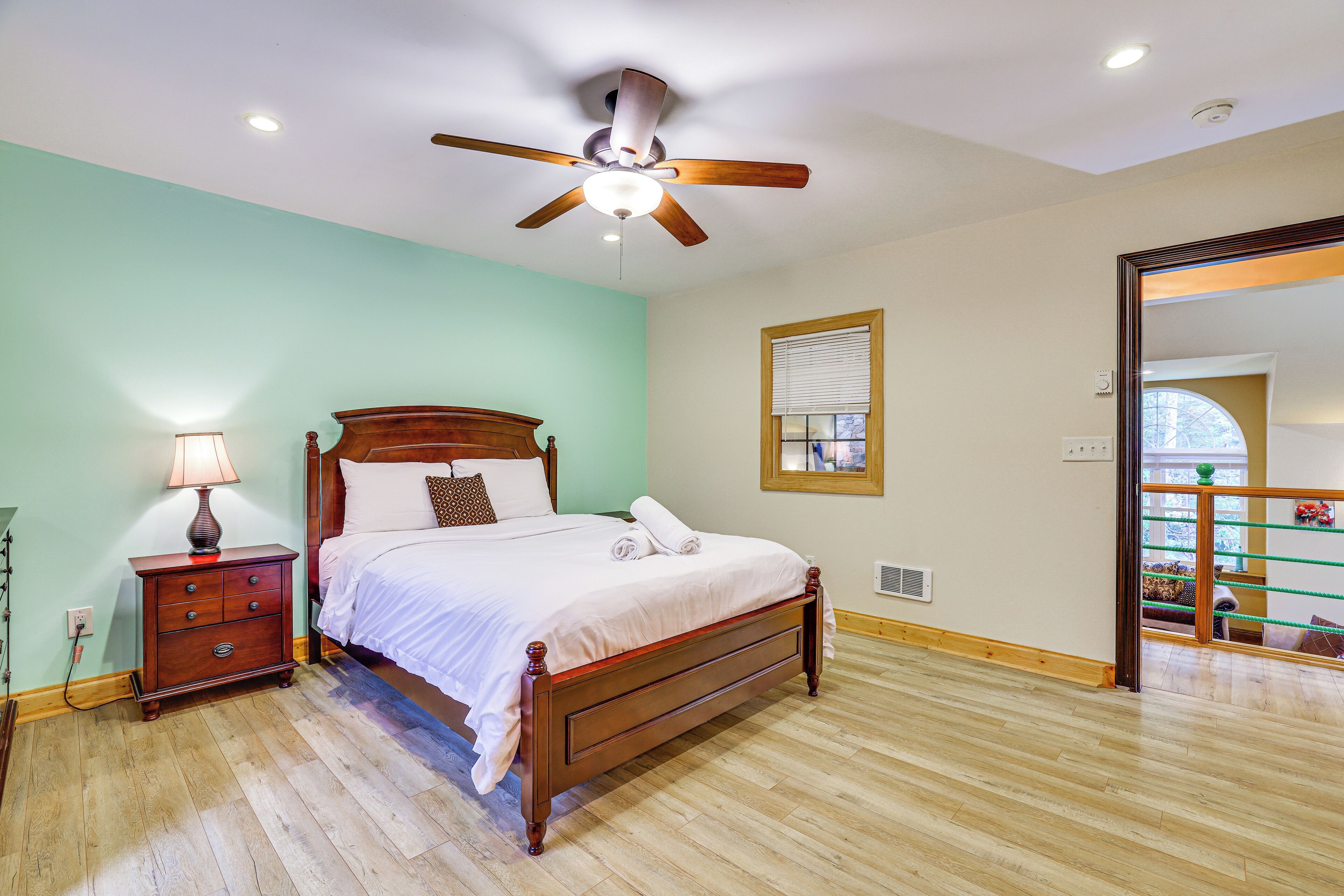 Un dormitorio con una cama de madera, una mesita de noche con lámpara, un ventilador de techo y una ventana con persianas.