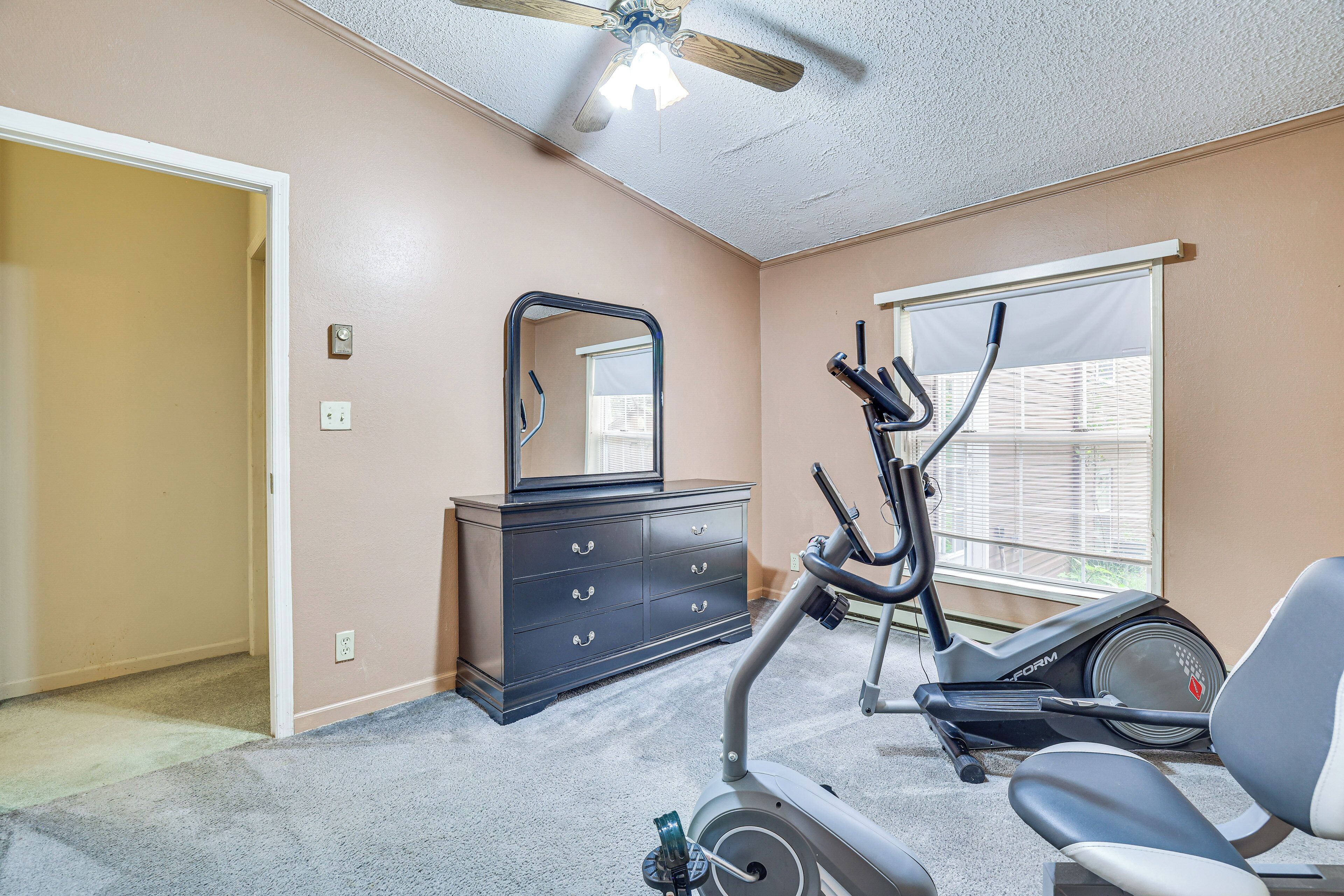 Une chambre avec des murs de couleur beige, une commode sombre, un miroir, un vélo d’appartement et un ventilateur de plafond.