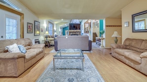 A spacious living room with two beige sofas, a glass coffee table, and a patterned rug. A kitchen area is visible in the background with a fireplace.