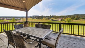 A covered outdoor area with a wooden table and wicker chairs, overlooking a green landscape with houses and trees. - Deck & Hot Tub: Group Golf Retreat in Sturgis (Sturgis)