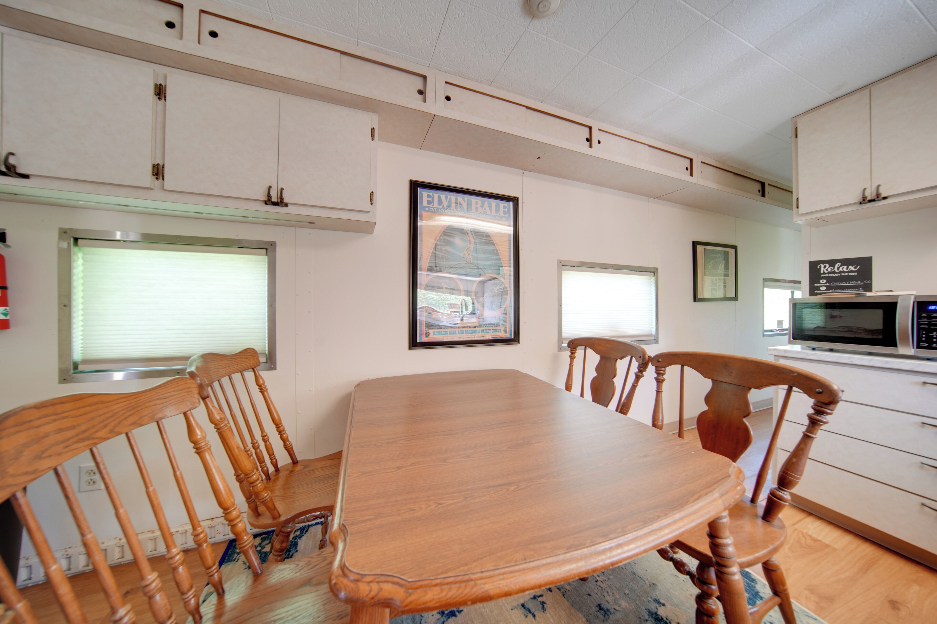 A dining area with a wooden table and chairs, a framed picture on the wall, and a microwave in the background.