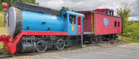 A vintage steam locomotive with a red and blue caboose, parked on a track with a gravel bed and a paved area beside it.