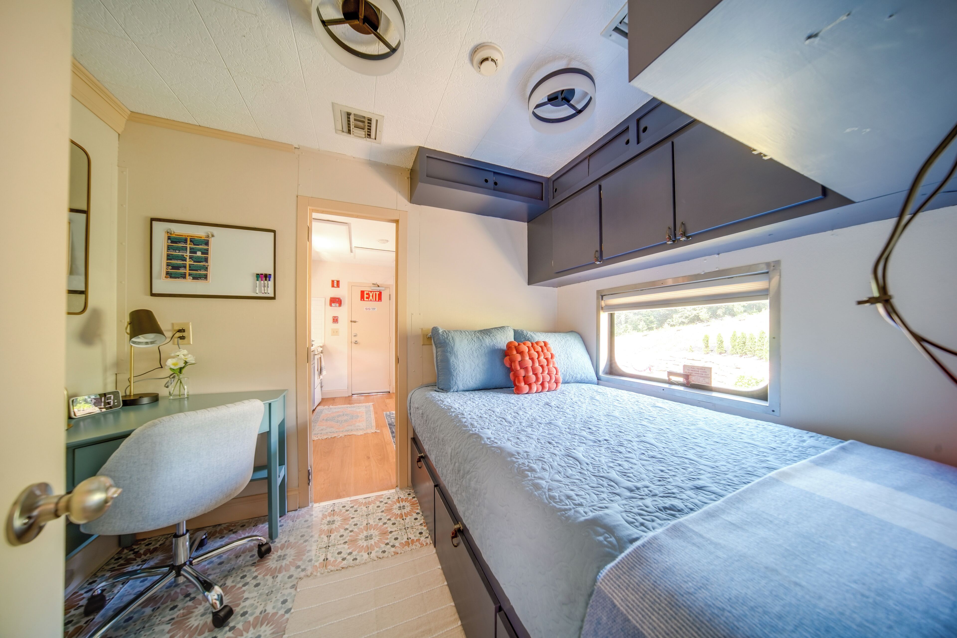 A compact bedroom with a bed, a desk, and a chair. There is a window providing natural light and a door leading to another room.