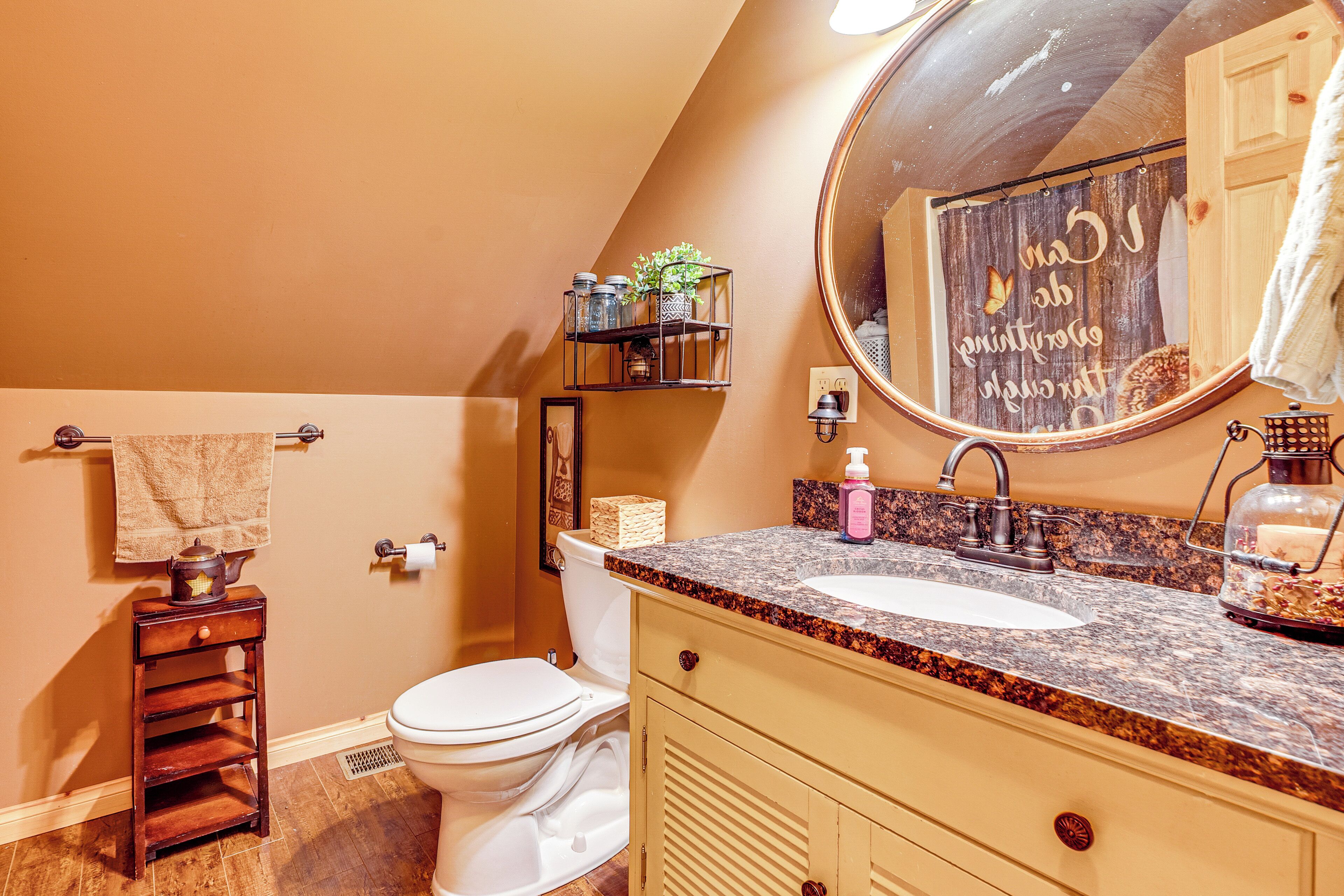 A bathroom with a white toilet, a sink with a granite countertop, a mirror, and a shelf with toiletries.