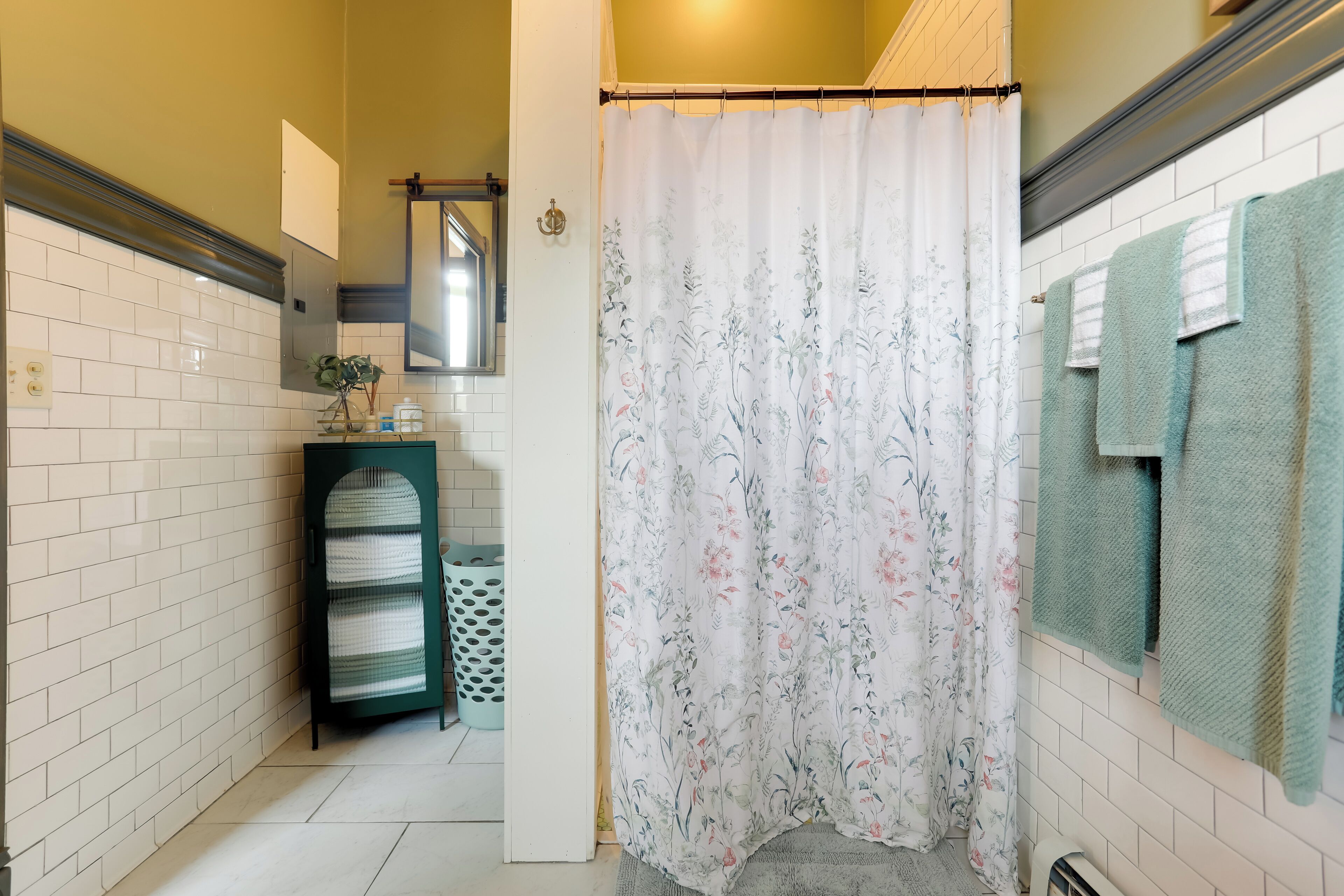 Un baño con paredes de azulejos blancos, un perchero verde, una cortina de ducha con motivos florales y una toalla verde colgada en la pared.