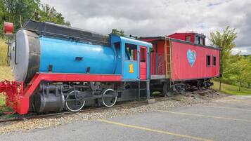 A vintage steam locomotive with a red and blue caboose, parked on a track with a gravel bed and a paved area beside it.